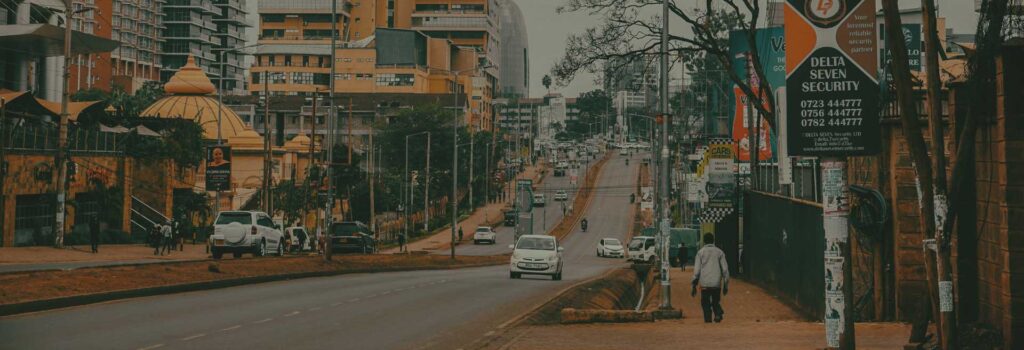Nairobi street scene with cars on a main road, city buildings, roadside billboards, and a person walking along the pavement.