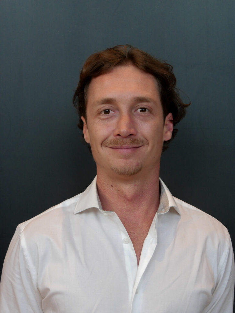 Smiling man in a white shirt against a dark blue background, conveying a professional and approachable demeanor.
