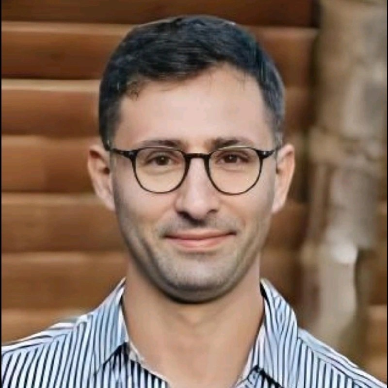A professional man with glasses and a striped shirt smiles against a wooden background, representing Yalelo Uganda.